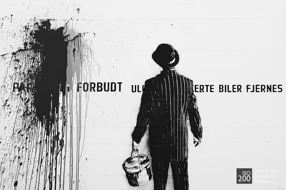 Stencil and freehand graffiti of a man in a suit with a bowler hat painting graffiti on a wall in the docks of Stavanger, Norway.